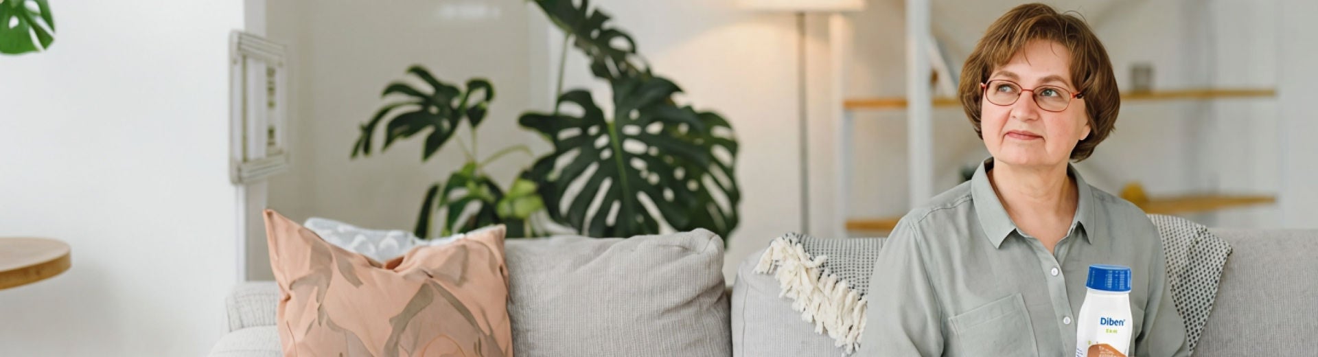 A woman sits on a sofa at home and gently smiles whilst holding a Diben Apricot-Peach flavoured Drink from Fresubin to help support her nutritional needs and diabetes. 