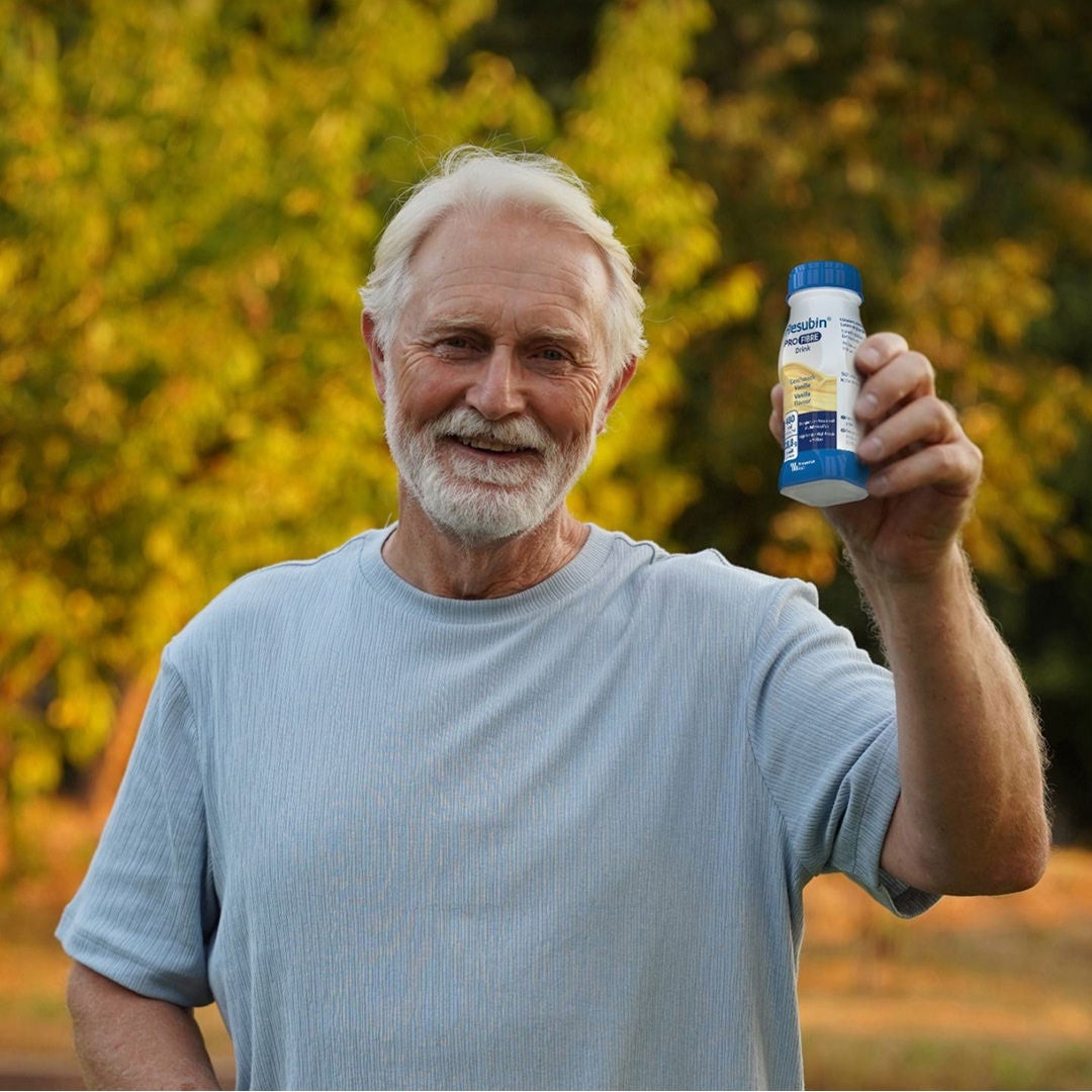 Un hombre de edad avanzada, de cabello canoso y vestido con una camiseta gris, sostiene una botella de PRO Drink de Fresubin con sabor a vainilla, un producto que ofrece un apoyo nutricional de alta densidad para un envejecimiento saludable. 