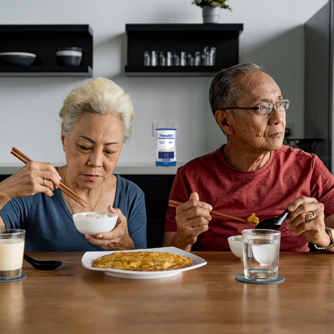 Twee personen eten aan een tafel in een keukenomgeving. Op het aanrecht staat een verpakking van Fresubin Protein Powder.