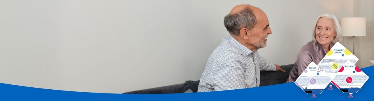 Un hombre y una mujer de edad avanzada, sentados en un sofá, se sonríen mutuamente, conscientes de que cuentan con el respaldo de la gama de alimentos para alimentación por sonda de Fresubin, que incluye Frebini ORIGINAL FIBRE, Fresubin ENERGY y Supportan Tube Feeds. 