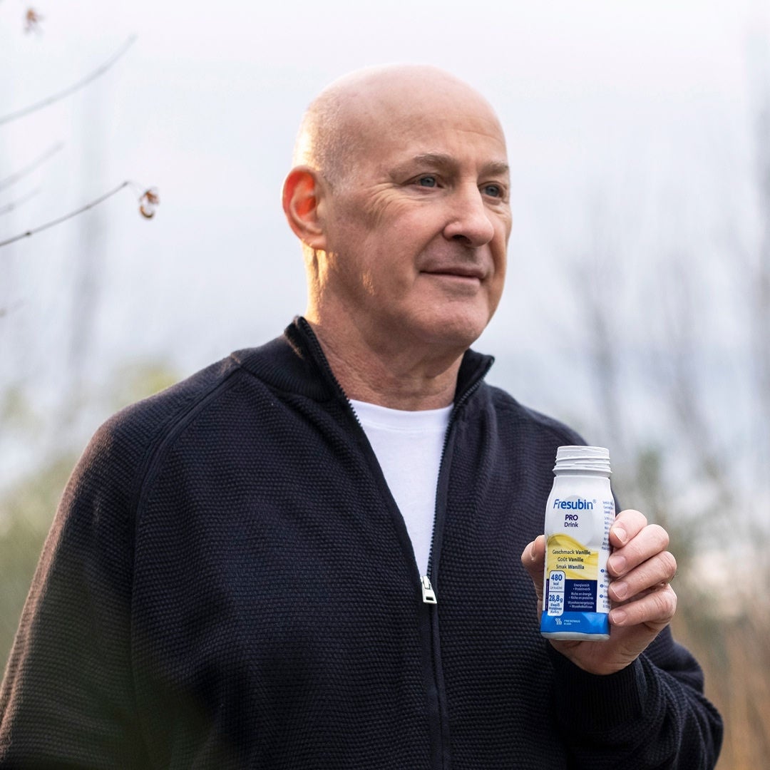 An older man with a shaved head wearing a black zip-up hoodie holds a bottle of Fresubin’s Vanilla-flavoured PRO Drink that offers high density nutritional support for healthy ageing. 