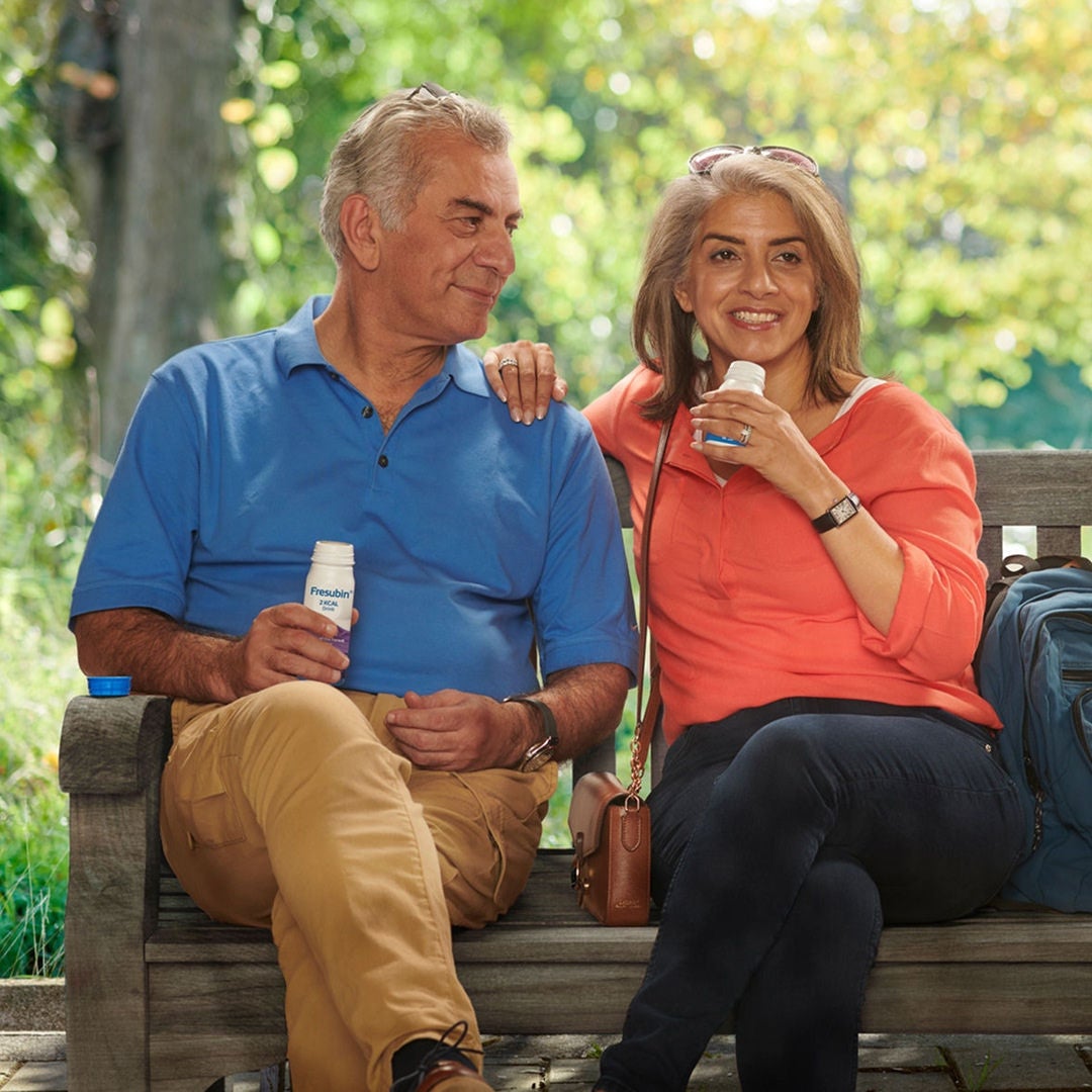 Una pareja está sentada en un banco de madera disfrutando de Fresubin 2KCAL Drink, un suplemento nutricional oral rico en energía y proteínas, indicado para cubrir un mayor requerimiento energético. 
