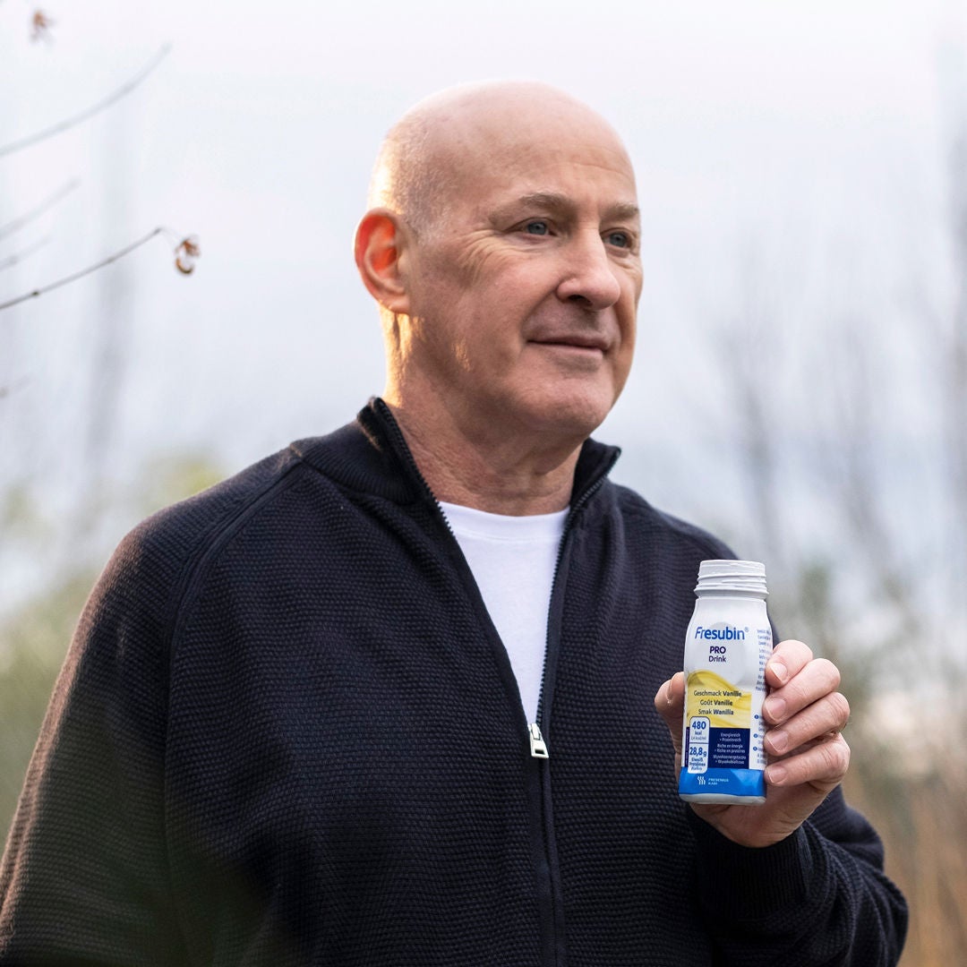 Um homem de idade avançada, com a cabeça rapada e vestindo um casaco com capuz preto com fecho, segura uma garrafa de PRO Drink da Fresubin com sabor a baunilha, que proporciona um apoio nutricional de alta densidade para um envelhecimento saudável. 
