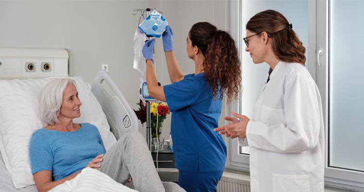 Das Bild zeigt eine ältere Frau in einem Krankenhausbett, während eine Pflegekraft Fresubin Sondennahrung vorbereitet und eine Ärztin das Geschehen beaufsichtigt. 