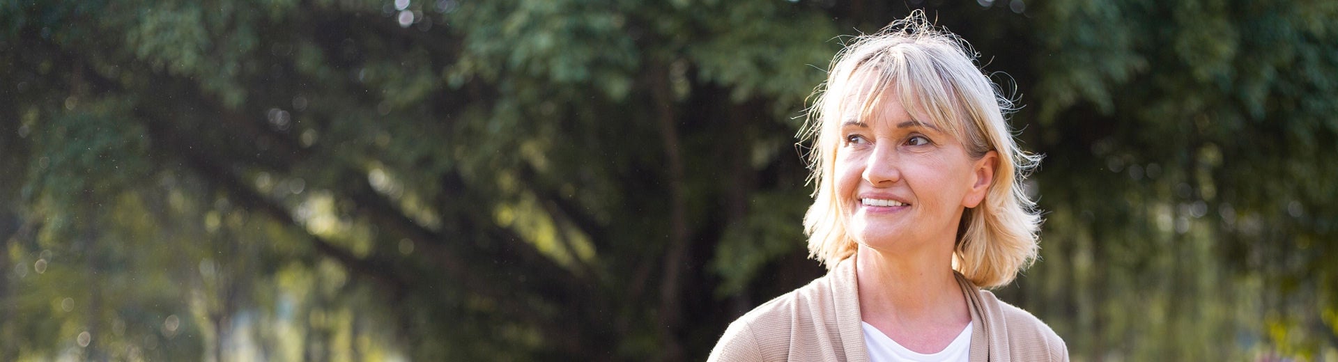 Una mujer rubia sonríe tranquilamente al aire libre en un parque bañado por el sol, lo que representa a una paciente que lleva una vida plena a pesar de padecer una enfermedad renal crónica gracias al apoyo nutricional que ofrece la gama Fresubin 