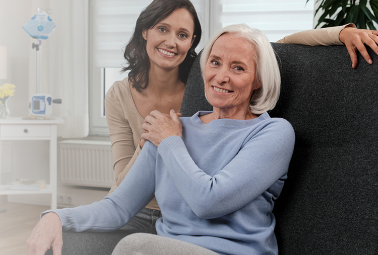 Lächelnde ältere Frau, die auf einem Stuhl sitzt, mit einer jüngeren Frau, die neben ihr steht, beide zeigen Wärme und Zuneigung in einer hellen Innenumgebung.