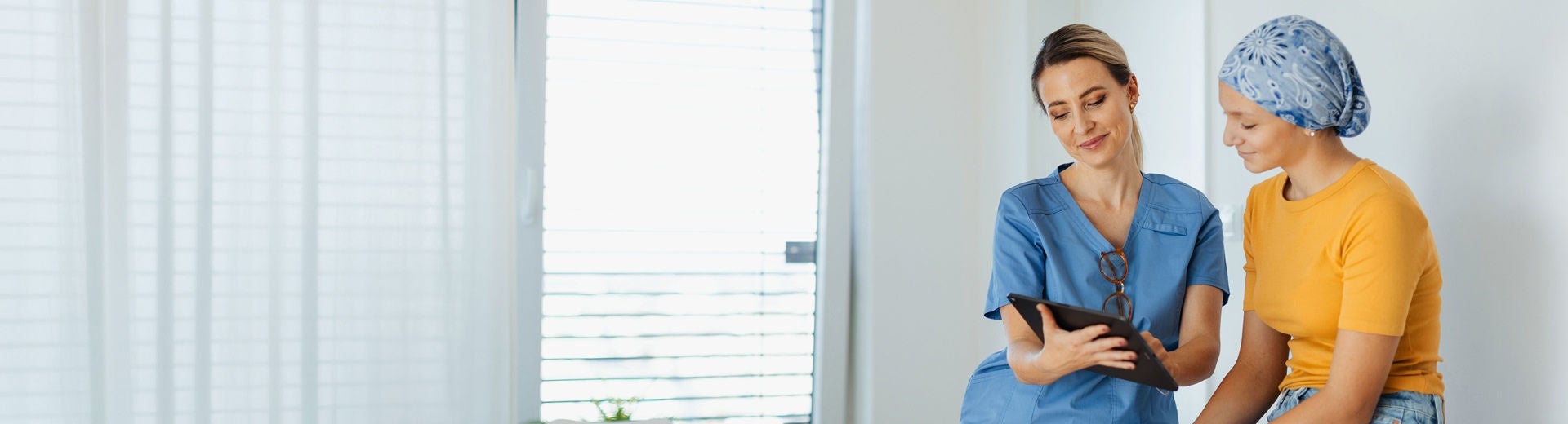 Un profesional de la salud, vestido con una bata azul, revisa en una tableta la información nutricional y los consejos destinados a una joven paciente que lleva un pañuelo azul en la cabeza, incluyendo cómo Fresubin puede ayudar a controlar la nutrición a lo largo del proceso de tratamiento del cáncer. 