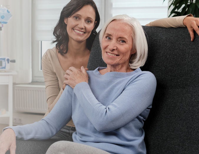 Smiling elderly woman sitting in a chair with a younger woman standing beside her, both showing warmth and affection in a bright indoor setting.