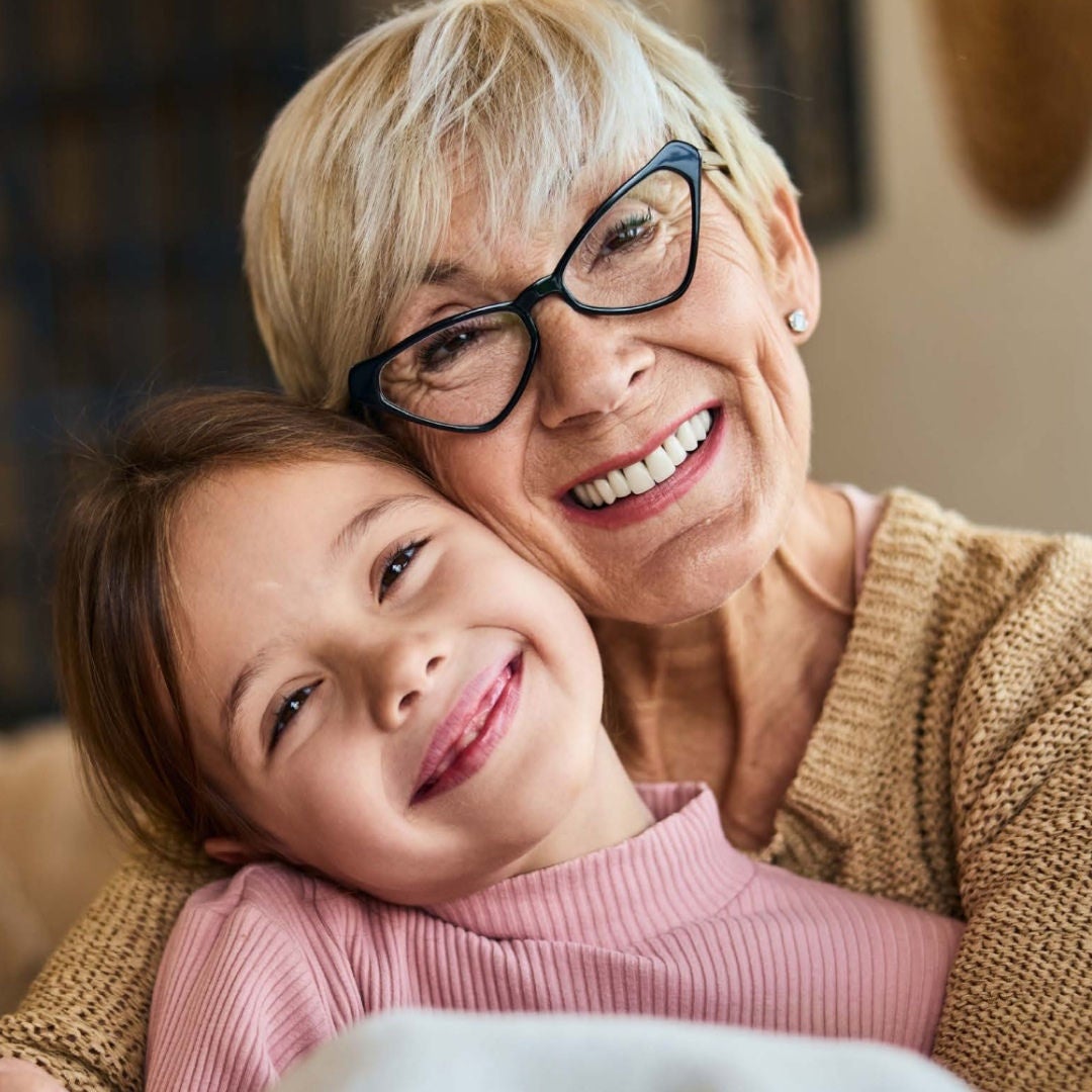 An older woman with short grey hair and glasses, and a small girl smile warmly while sharing an embrace 