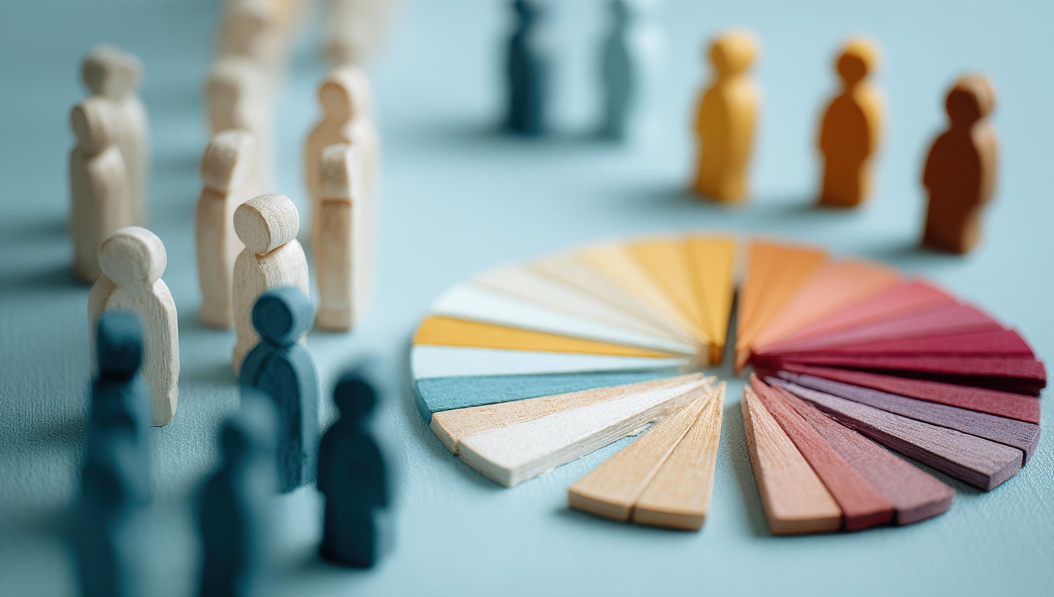 Holzfiguren in verschiedenen Farben stehen in Gruppen auf einer hellblauen Fläche. Daneben liegt ein fächerförmig angeordnetes Farbdiagramm aus Holzleisten, das unterschiedliche Farbtöne von Beige über Gelb bis Rot und Blau zeigt. Das Bild symbolisiert Vielfalt, Gruppenbildung und unterschiedliche demografische oder gesellschaftliche Zusammenhänge.