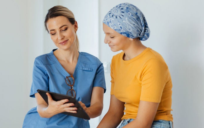 Um profissional de saúde, vestindo uma blusa cirúrgica azul, analisa informações e orientações nutricionais em um tablet com uma jovem paciente que usa um lenço azul na cabeça, incluindo como o Fresubin pode ajudar no controle da nutrição ao longo do tratamento do câncer. 