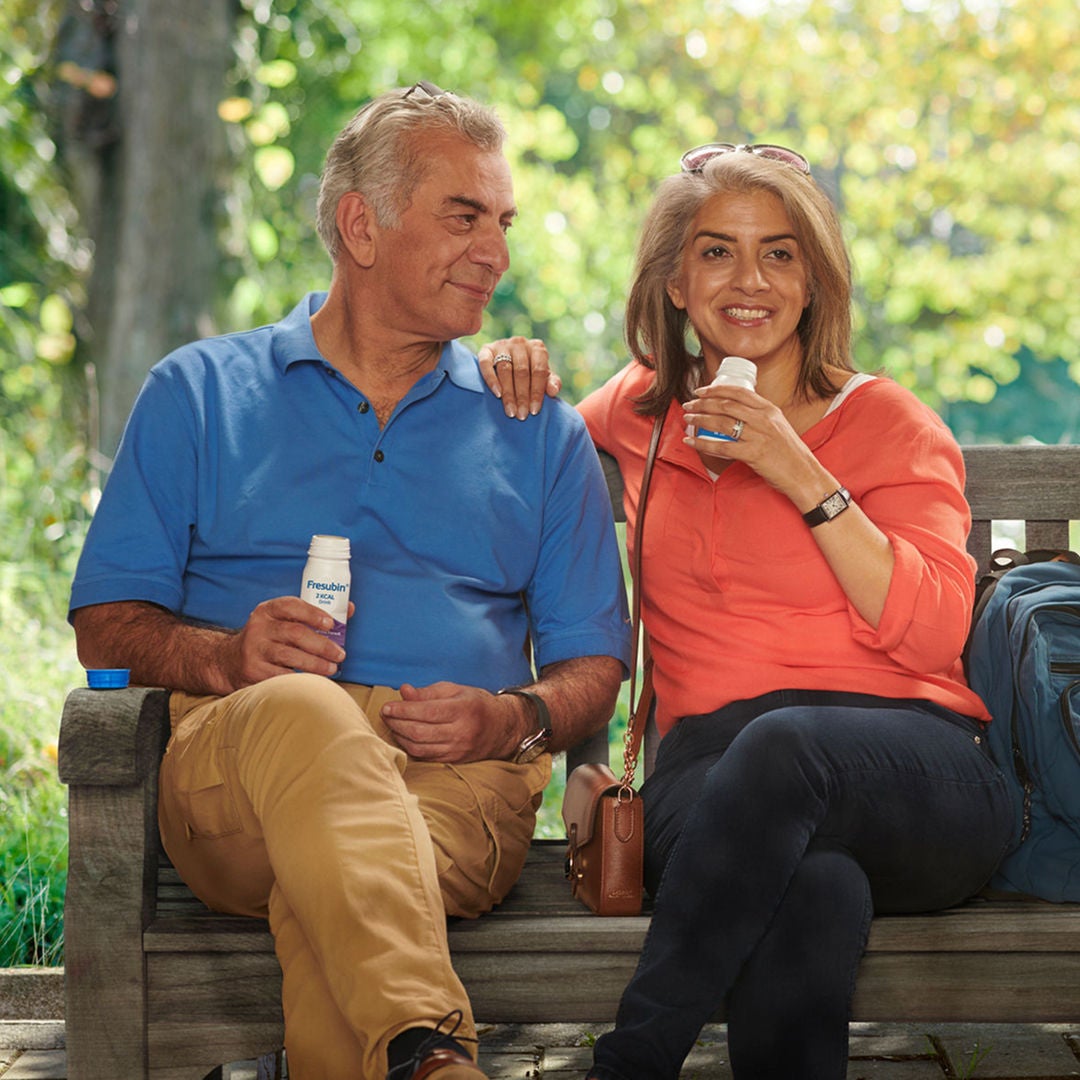 Um casal está sentado em um banco de madeira, saboreando a bebida Fresubin 2KCAL, um suplemento nutricional oral rico em energia e proteínas, indicado para atender a necessidades energéticas elevadas. 