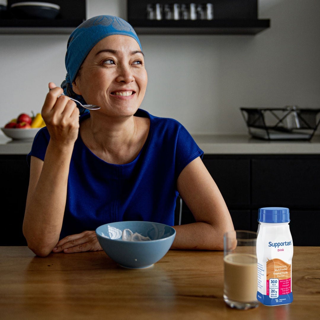 O pacientă cu cancer, purtând o eșarfă albastră și o bluză, zâmbește în timp ce savurează micul dejun și băutura Fresubin Supportan cu aromă de cappuccino, care o ajută să-și acopere nevoile nutriționale 