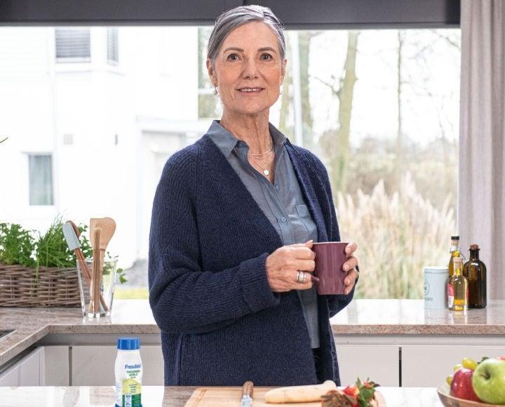 A lady holding a purple mug enjoys a Vanilla-flavoured Fresubin Thickened Level 2 Drink in her kitchen as part of her daily nutritional intake and dysphagia management.  