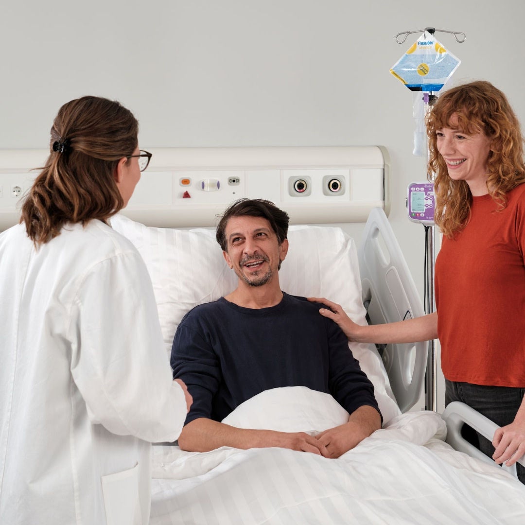 Un hombre sonríe desde su cama de hospital a su profesional de la salud, mientras recibe apoyo nutricional gracias a la gama de alimentación por sonda HP ENERGY de Fresubin. 