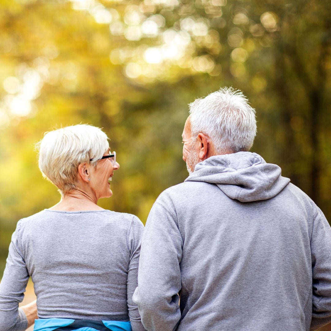 An older couple turn to smile at each other as they walk away through a forest, knowing their nutritional needs are supported by Fresubin's product range, including Fresubin Protein Shot. 