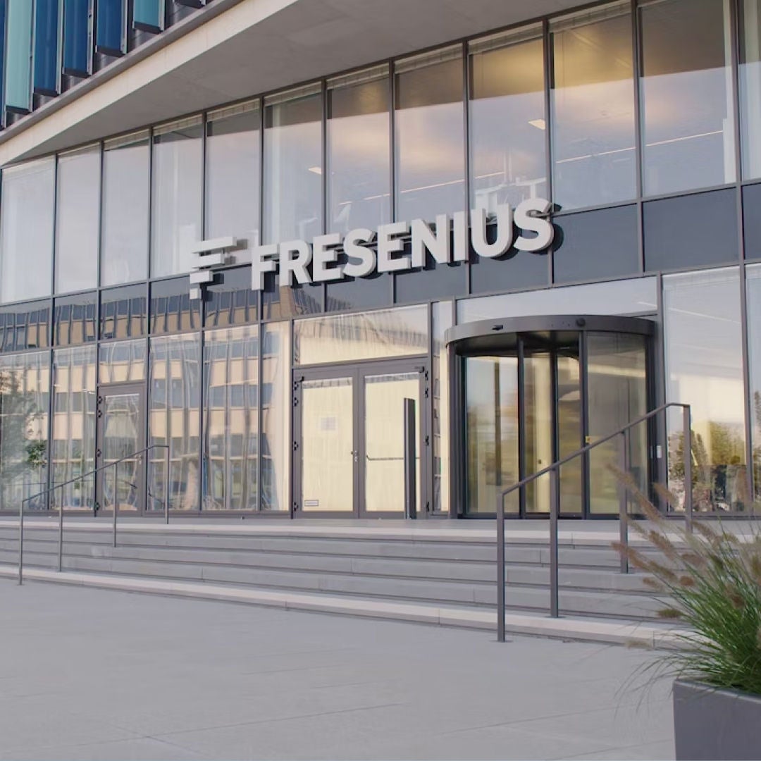 Front view of a modern, grey Fresenius office building with glass front and stairs leading up to the entrance. 