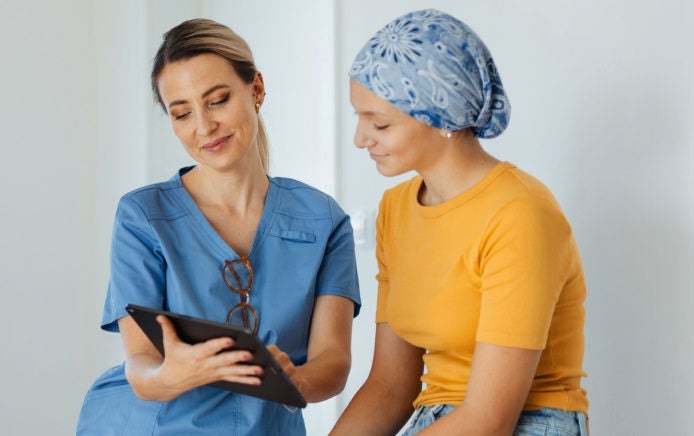Un profesional de la salud, vestido con un uniforme azul, revisa en una tableta información y consejos nutricionales con una paciente joven que lleva un pañuelo azul en la cabeza, incluyendo cómo Fresubin puede ayudar a controlar la nutrición a lo largo del proceso de tratamiento del cáncer. 