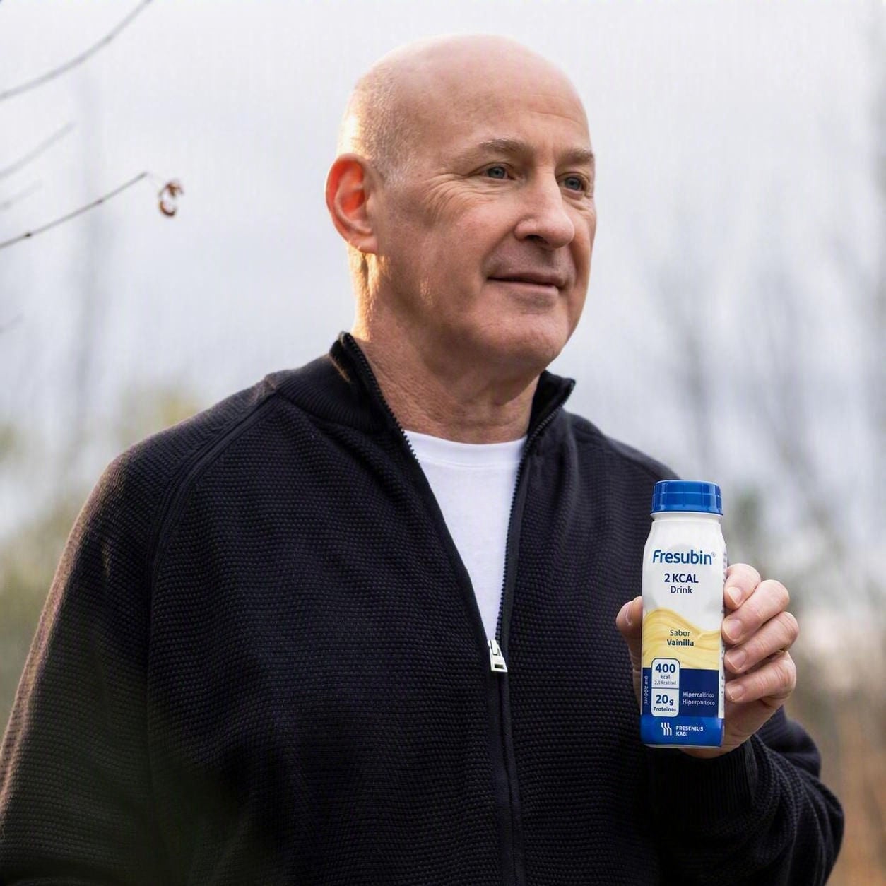 Un hombre de edad avanzada, con la cabeza rapada y vestido con una sudadera negra con capucha y cremallera, sostiene una botella de PRO Drink de Fresubin con sabor a vainilla, un producto que ofrece un apoyo nutricional de alta densidad para un envejecimiento saludable. 