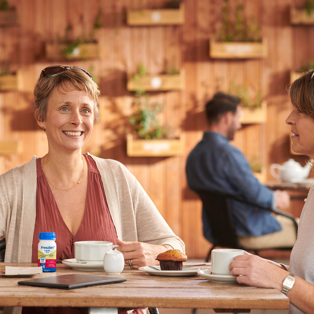 Persoon zit aan tafel met een kop koffie en een flesje Fresubin Protein Energy Drink op een dienblad.
