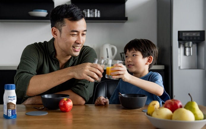 Un hombre sonriente está sentado junto a un niño sonriente en la barra de la cocina, mientras disfrutan del desayuno con una bebida Fresubin 2KCAL con sabor a chocolate. 