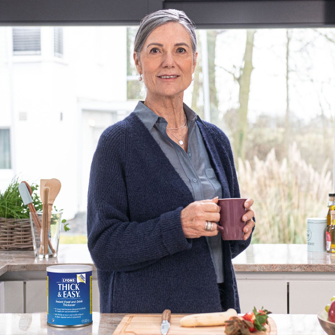 Una mujer que sostiene una taza morada disfruta de una bebida Fresubin Nivel 2 con sabor a vainilla en su cocina, como parte de su ingesta nutricional diaria y del tratamiento de su disfagia. 