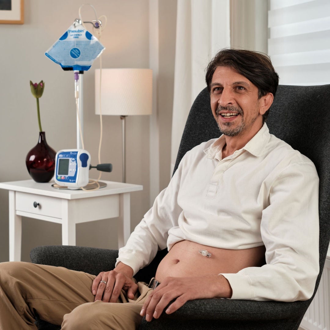A man with a tube feeding port on his stomach, smiles as he sits in a chair at home with Fresubin HP ENERGY FIBRE tube feed next to him to support his nutritional needs. 