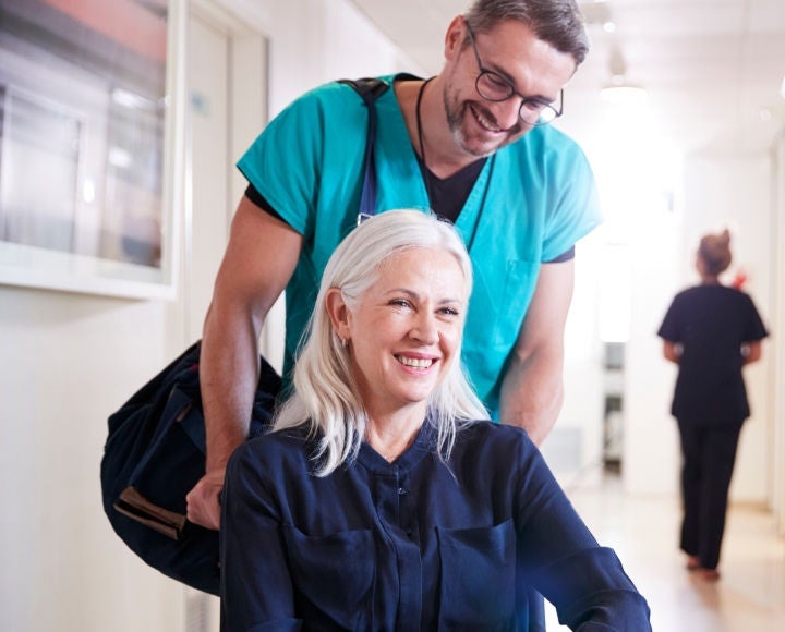 A healthcare professional in light blue scrubs pushes a woman in a wheelchair who smiles knowing she is supported by Fresubin's nutritional supplement range. 