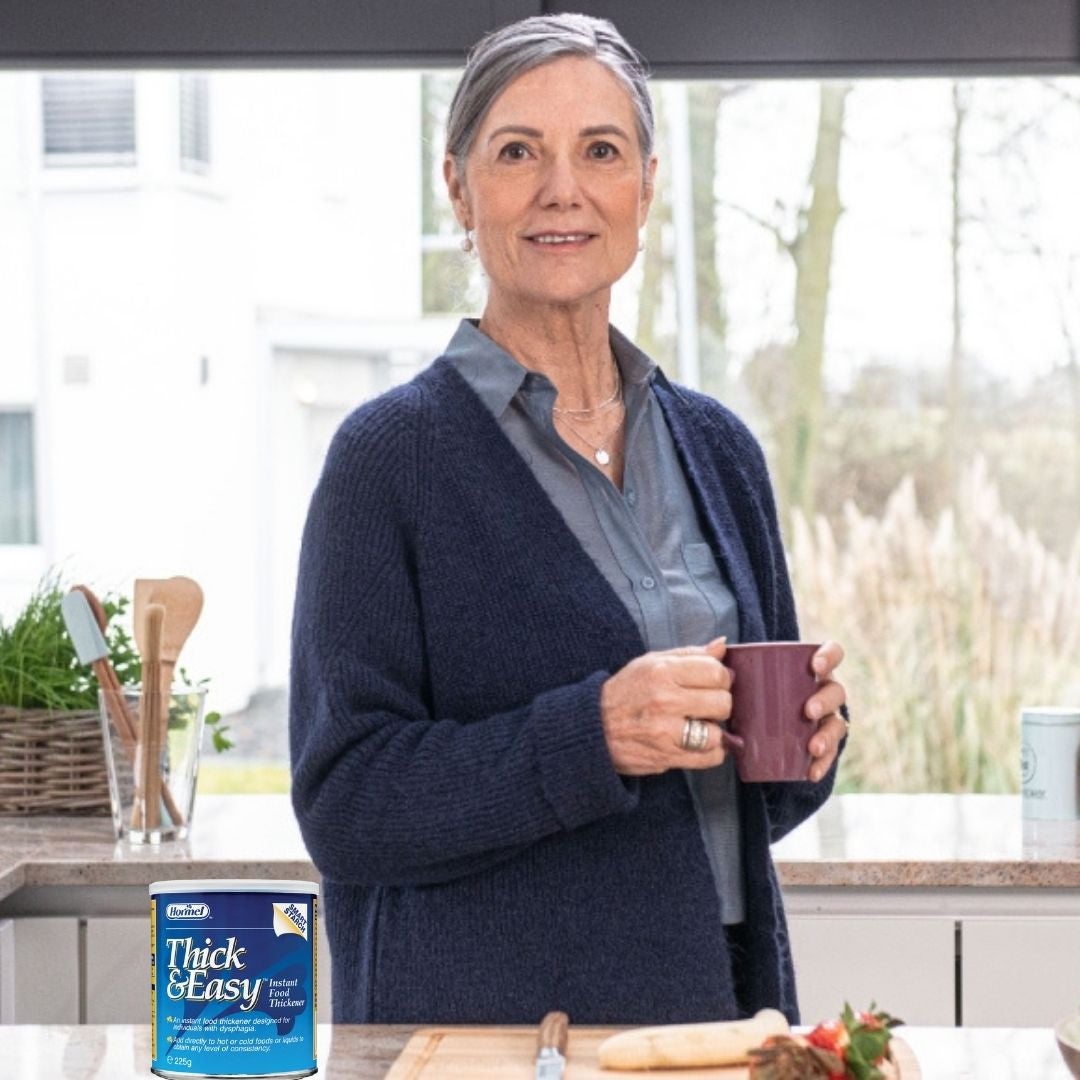 Una mujer que sostiene una taza morada disfruta de una bebida Fresubin espesada de nivel 2 con sabor a vainilla en su cocina, como parte de su ingesta nutricional diaria y del tratamiento de su disfagia.  