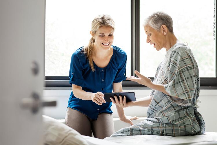 Eine ältere Patientin im Krankenhaushemd hält ein Tablet in der Hand und spricht mit einer jüngeren, lächelnden Pflegekraft in blauer Kleidung - verlinkt zu Fresenius Kabi Homecare