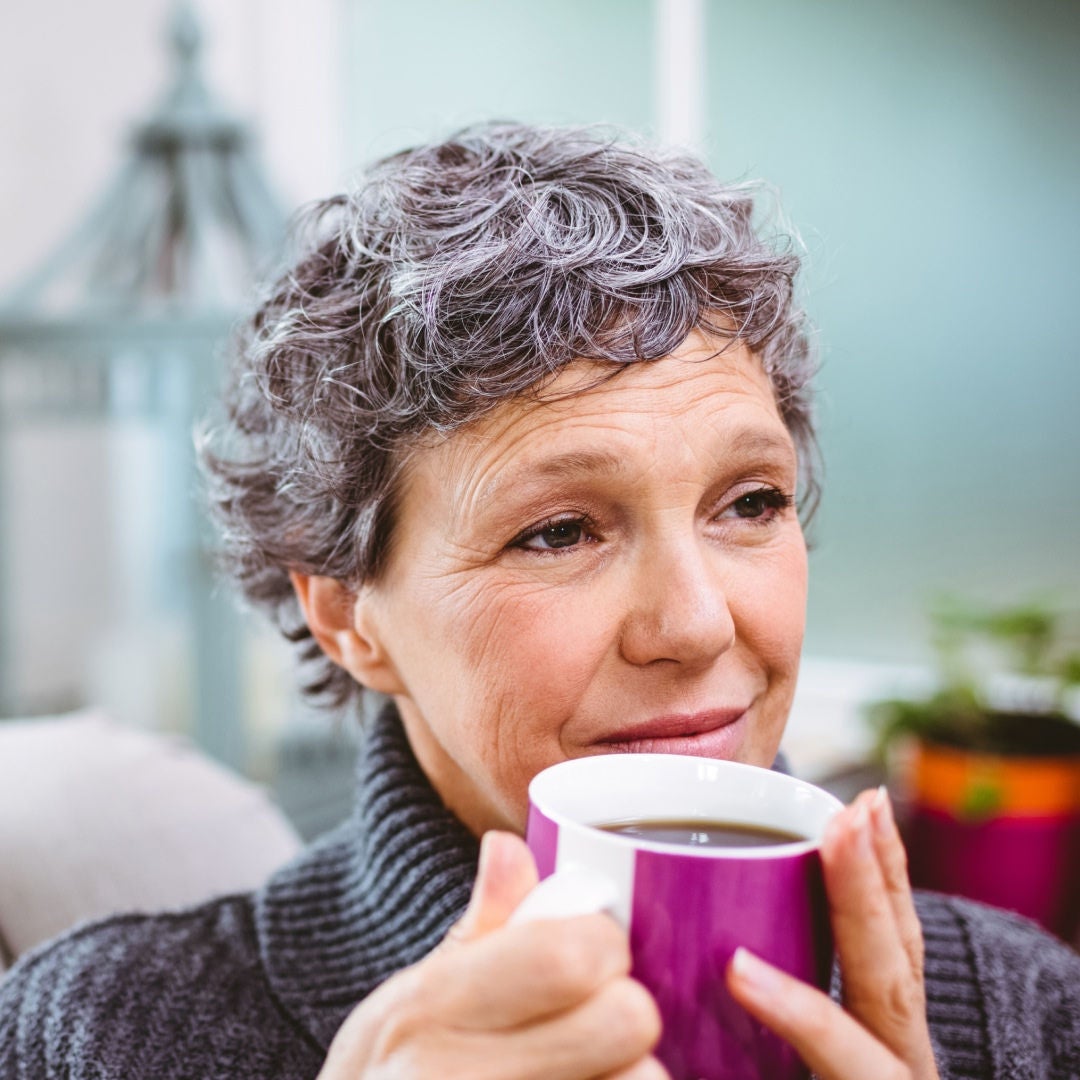 Una mujer de cabello corto, rizado y canoso sonríe con dulzura mientras sostiene una taza de café morada, consciente de que cuenta con el respaldo de la gama de suplementos nutricionales de Fresubin 