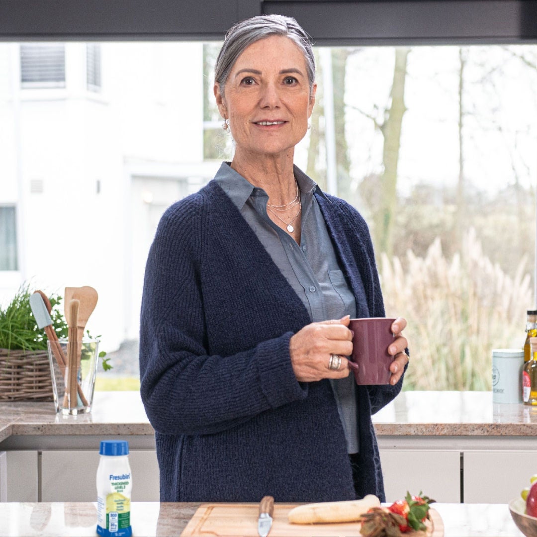 Una mujer que sostiene una taza morada disfruta de una bebida Fresubin Nivel 2 con sabor a vainilla en su cocina, como parte de su ingesta nutricional diaria y del tratamiento de su disfagia. 