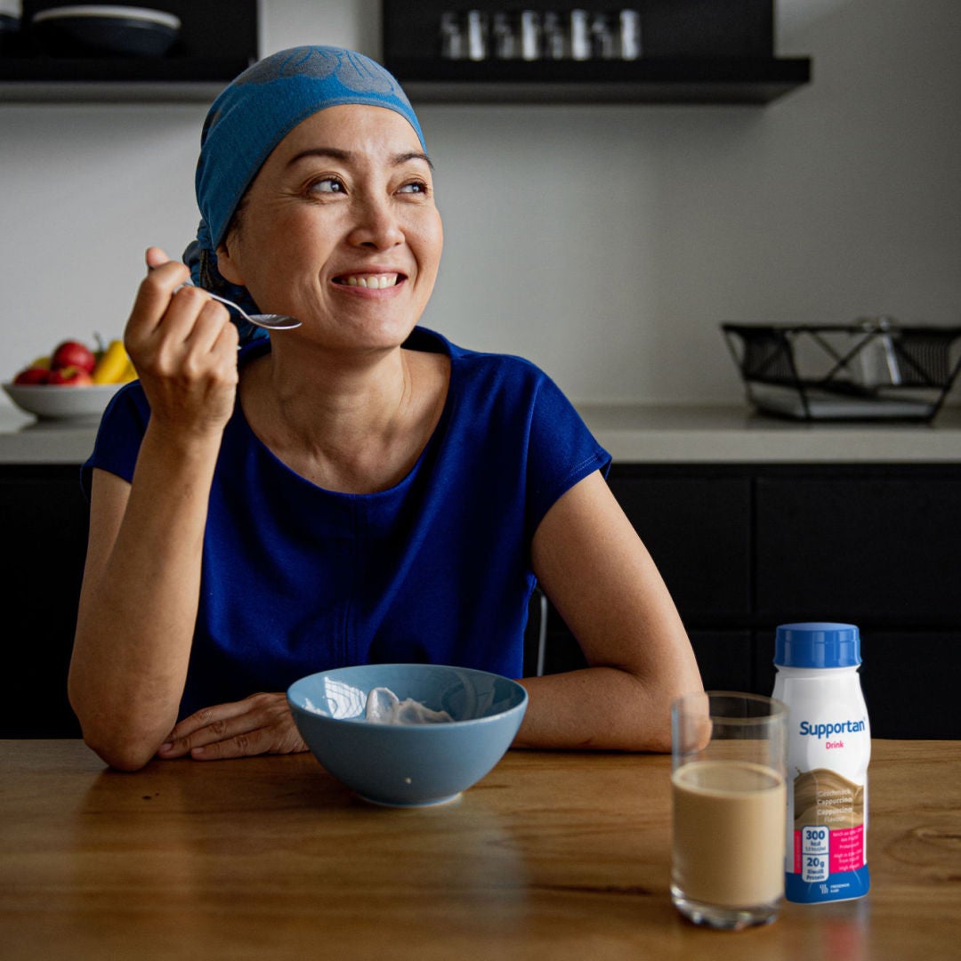 Uma doente com cancro, com um lenço azul na cabeça e uma blusa, sorri enquanto saboreia o pequeno-almoço e a bebida Supportan da Fresubin com sabor a cappuccino, que a ajuda a satisfazer as suas necessidades nutricionais 