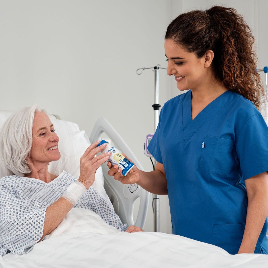 Una mujer de edad avanzada, recostada en una cama de hospital, le sonríe a su profesional de la salud, quien le está entregando una bebida Fresubin PRO con sabor a vainilla. 