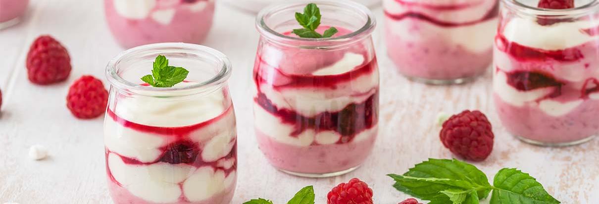 Mehrere Gläser mit Himbeer Trifle und daneben Himbeerdekoration auf einer weißen Holzoberfläche.