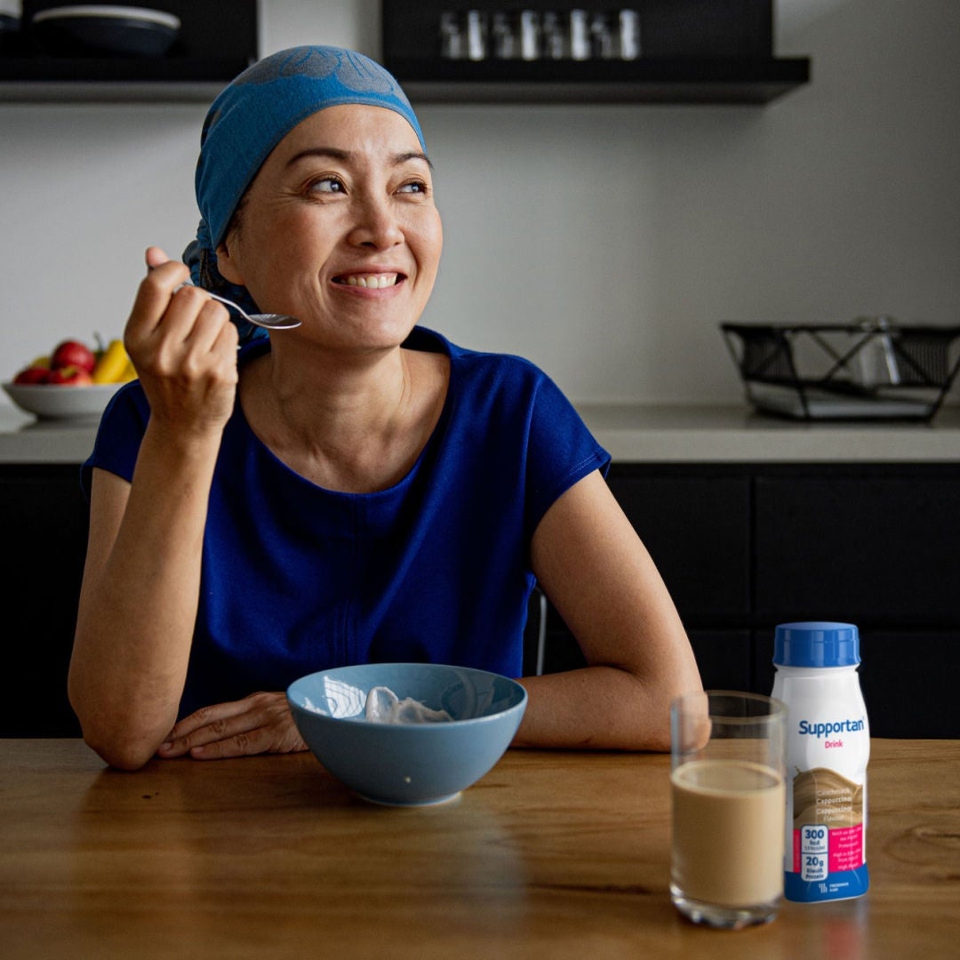 Una paciente de cáncer, con un pañuelo azul en la cabeza y una blusa, sonríe mientras disfruta de su desayuno y de la bebida Supportan de Fresubin con sabor a capuchino, que le ayuda a cubrir sus necesidades nutricionales 