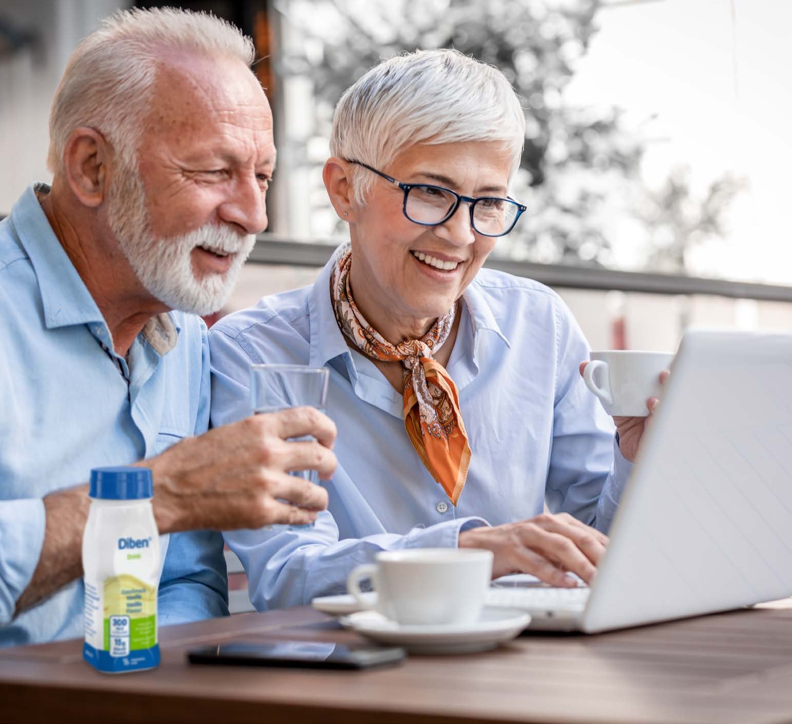 An older man and woman sit together at a laptop whilst enjoying a Diben Vanilla-flavoured Drink, designed to support the nutritional needs of those with diabetes. 