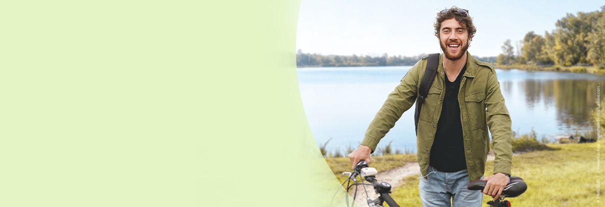 Mann steht vor einem See mit seinem Fahrrad in der Hand und schaut freundlich in die Kamera