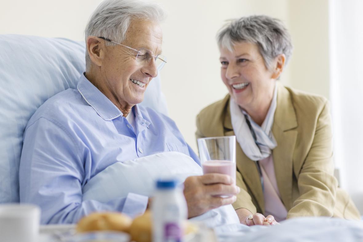 Älterer Patient im Krankenhausbett hält ein Glas mit Fresubin in der Hand. Seine Partnerin sitzt als Besucherin neben ihm.