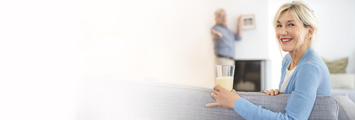 Eine Frau, lehnend über dem Sofa, hält ein Glas mit Fresubin Trinknahrung, für besondere medizinische Zwecke (bilanzierte Diät).